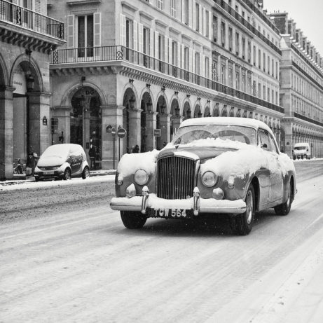 A vintage car covered in neige drives down a snow-covered street lined with elegant buildings with archways and balconies. Snow continues to fall, adding to the wintry atmosphere of this quiet dimanche morning. Other vehicles are parked along the street, also dusted with snow.