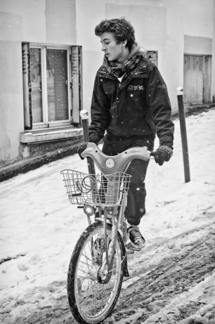 A person wearing a winter jacket, scarf, and gloves rides a bicycle on a snowy street. Snowflakes are falling, and the street is covered in neige. In the background, there are buildings with shuttered windows and a partially visible door, capturing the quiet beauty of a typical dimanche afternoon.