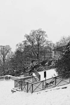 A black and white photo of a snowy scene in a Paris park with bare trees and a snow-covered pathway. A metal fence lines the path. In the background, neige-laden trees partially obscure a row of buildings. Snow blankets the ground and tops the fence.