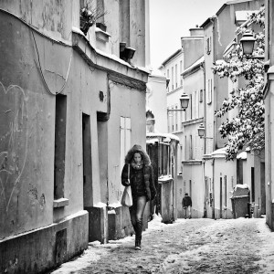 Black and white image of a snow-covered urban street with buildings on both sides. A woman with curly hair, wearing a winter jacket and boots, walks toward the camera carrying a bag. It's dimanche, and another person is visible in the distance walking away through the neige.
