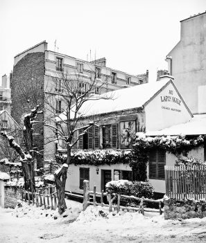 A black and white photograph captures a snow-covered street scene in hiver, featuring the historic cabaret "Au Lapin Agile" in Montmartre, Paris. Bare trees frame the building and a fence surrounds it, while apartment buildings stand visible in the background.