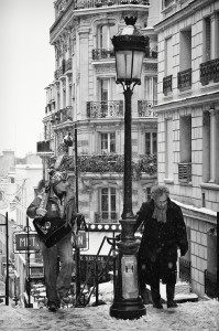 Black and white photo of a snowy street scene in Paris. A musician with a guitar and a heavily bundled older man are ascending a flight of stairs. The street, covered in neige, is lined with classic Parisian buildings, visible through falling snow and a vintage-style lamppost in the foreground.