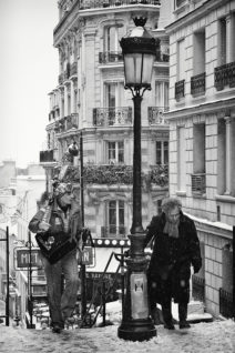 Black and white photo of a snowy street scene in Paris. A musician with a guitar and a heavily bundled older man are ascending a flight of stairs. The street, covered in neige, is lined with classic Parisian buildings, visible through falling snow and a vintage-style lamppost in the foreground.