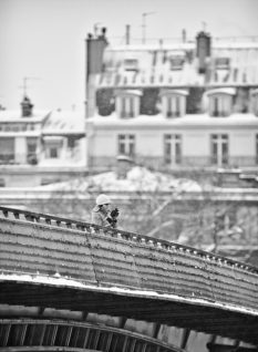 A lone person dressed in winter clothing stands on a snow-covered bridge, holding a camera. In the background, multi-story buildings with neige-laden rooftops are visible. The scene is monochromatic, emphasizing the cold, wintry atmosphere of a quiet dimanche.