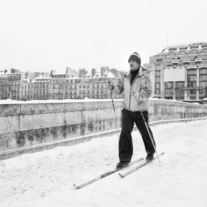 A skieur glides cross-country on a snowy city street near Pont-Neuf, with buildings and rooftops visible in the wintry background.
