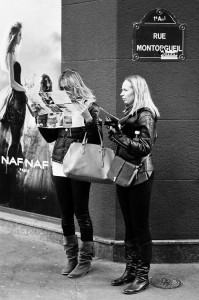 Two désorientées women in jackets and boots stand on a city sidewalk; one is reading a map while the other looks ahead holding a phone. Behind them, a fashion poster and a street sign reading “Rue Montorgueil” set the scene.