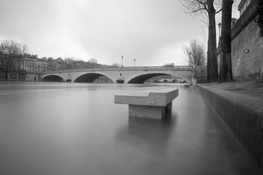 A serene black-and-white photograph of a bridge over a calm river. A long exposure captures the smooth water effect, with trees lining the riverbank and a submerged bench in the foreground. The sky is overcast, creating an almost "attention ça déborde" tranquil, reflective atmosphere.