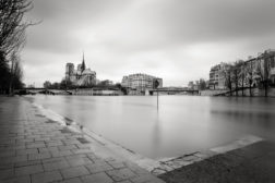 A black and white photograph of the Notre-Dame Cathedral in Paris, viewed from across the Seine River. The riverbank in the foreground is empty, and the sky above is overcast, creating a serene, reflective scene that demands attention.
