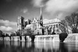 Black-and-white photo of Notre-Dame Cathedral in Paris, seen from across the River Seine. The gothic architecture features prominent spires, detailed façades, and intricate stonework. Trees line the riverbank, and a slight blur suggests movement in the water and clouds. Attention-catching artistry!