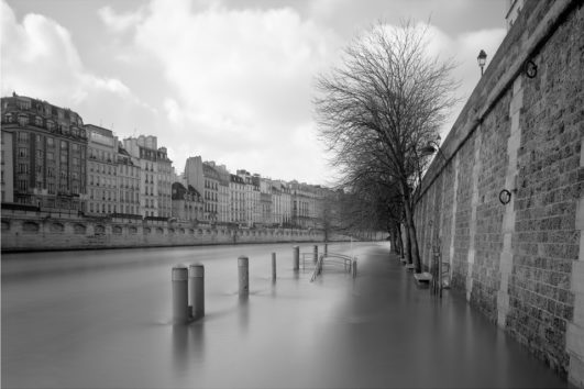 Black and white photo of a riverside promenade partially submerged by floodwaters. Leafless trees line the pathway next to a stone wall, with buildings visible in the background. Bollards and benches are nearly hidden by the floodwaters. Attention: Ça déborde under a cloudy sky overhead.