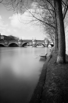 Black and white photo of a serene river with a stone bridge in the background. Bare trees line the walkway along the riverbank, and a bench sits overlooking the calm water. Attention is drawn to the tranquil scene with soft light and a misty, dreamy ambiance.