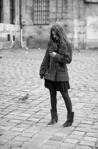 A belle with long hair, wearing a plaid jacket and boots, stands on a cobblestone street looking at her phone. Earphones hint at her musique, while an oiseau walks nearby. The photo is in black and white.