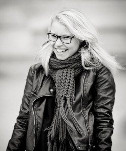 A smiling woman with light hair and glasses wears a scarf and a leather jacket. The photo is black and white and her hair is gently blown by the wind.