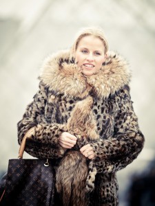 A woman smiles while wearing a leopard-print fur coat with a large collar, holding it closed and carrying a monogram designer handbag. The blurred background hints at the chic scene of LA Fashion Week.