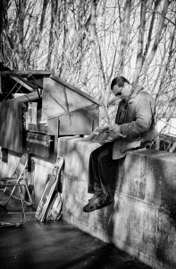 A man in a coat and glasses sits on a stone wall reading his journal du matin. An open street vendor cart and empty chairs are nearby, while bare tree branches fill the black and white background.