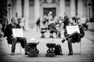 Two men sit facing each other on chairs outdoors, each holding a sign that reads "CONSEIL - ADVICE." A table is between them, and blurred people walk in the background. The image is in black and white.