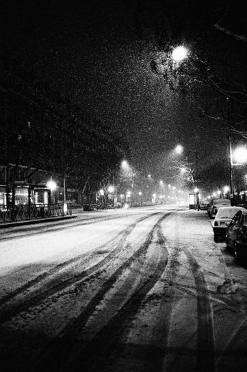 Black and white photo of a snowy, deserted street at nuit. Tire tracks can be seen on the neige-covered road, and streetlights line both sides, casting light through the falling snow. Parked cars and trees line the sidewalk, creating a serene, quiet atmosphere.