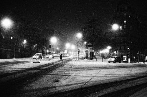 A black-and-white nighttime photograph of a snowy city street, illuminated by streetlights. Cars are parked along the side, and a few are driving. A lone person walks across the intersection. Snow covers the road and sidewalks, creating a serene winter atmosphere in this scene of nuit and neige.