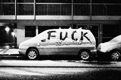 A car covered in neige is parked on a snowy street. The word "FUCK" is written in large letters on the snow covering the side of the car. The scene, captured in black and white, depicts the snowy weather and urban setting at nuit.