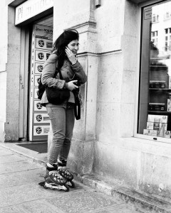 A patineuse souriant se tient sur un trottoir de la ville, tenant son visage d’une main tout en téléphonait. Elle porte un sac et se trouve près de l’entrée d’une pharmacie, avec des étagères et des enseignes en arrière-plan.