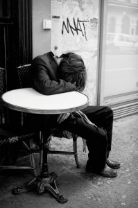 A person in a suit sits slouched at a round outdoor table, head resting on their arm and face obscured, revealing the Paris hidden side in this black and white scene with graffiti on the wall and a window in the background.