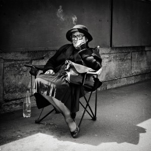 An elderly woman in dark clothing, hat, and large glasses sits on a folding chair by a wall along the quais de Paris, holding a cigarette. A bag rests on her lap and a bottle of water is at her feet. The black-and-white scene evokes vieux Paris.