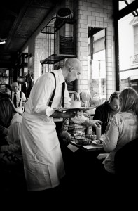 A waiter in a white apron and suspenders serves petit-déjeuner and drinks to a group of diners at a busy, stylish restaurant with tiled walls and large windows. The atmosphere is lively and bustling.