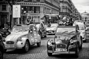 Two vintage Citroën 2CV cars with open tops cruise through an old school Paris traffic jam, surrounded by historic buildings. Two people in the cars snap photos and enjoy the vibrant city scene.
