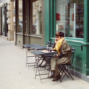 A person with short hair and an écharpe jaune sits alone at an outdoor café table, talking on the phone, surrounded by empty tables and city buildings in the background.