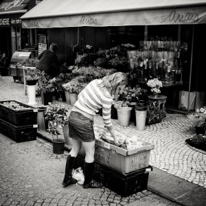 A woman in a striped shirt, skirt, and boots bends over a crate of goods outside a flower shop on a cobblestone street. The shop, with “Anaïs” on the awning, displays flowers and plants in buckets.