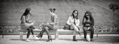 Quatre femmes sont assises sur un banc en plein air, discutant et mangeant. L'image en noir et blanc montre une pelouse à l'arrière-plan et un chemin de gravier au premier plan.