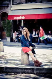 A young woman with long hair sits on a bollard by the street, wearing a red dress, dark jacket, and brown boots. Behind her is a café with a red awning and people at outdoor tables—Bientôt l'été fills the air with anticipation.