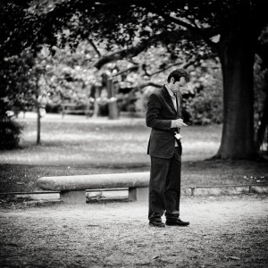 A man in a suit stands alone in a jardin public, looking down at his phone as if engaged in rédaction virtuelle. A stone bench and large trees are in the background. The image is in black and white.