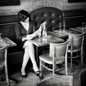 A woman with short dark hair sits alone at a restaurant table, reading a newspaper. Dressed in a dark outfit and high heels, she waits amid empty glasses and a bottle of water—a black and white scene capturing anticipation before a première rencontre.