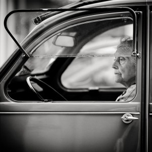 Black and white photo of an elderly woman sitting in the driver’s seat of a vintage car, looking thoughtfully out the window, as if recalling un temps she might have been moins de 20 ans. The car door and steering wheel are visible in the foreground.