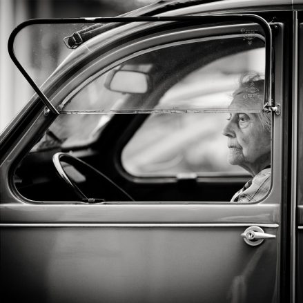 Black and white photo of an elderly woman sitting in the driver’s seat of a vintage car, looking thoughtfully out the window, as if recalling un temps she might have been moins de 20 ans. The car door and steering wheel are visible in the foreground.