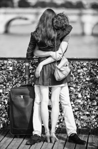 Les amoureux du pont stand embracing on a bridge covered with love locks. Seen from behind, they have a suitcase beside them, with a blurred river and another bridge in the background.