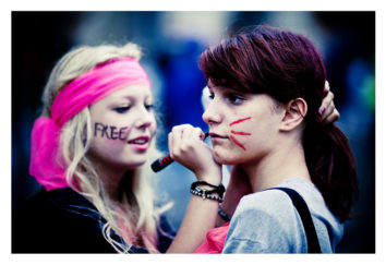 Two young women are standing close together at the 2013 Techno Parade. The woman on the left is wearing a pink headband and has "FREE" written on her cheek. She is using a marker to draw red lines on the right cheek of the woman with dark hair, celebrating the 15ème édition of this vibrant event.