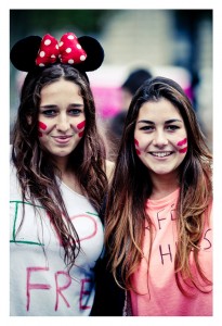 Two young women smiling, one wearing a black Mickey Mouse headband with a red polka-dot bow. Both have red streaks painted on their cheeks and are dressed casually, with "Techno Parade 2013" handwritten on their shirts. The background is blurred, focusing attention on the lively scene at the 15ème édition.