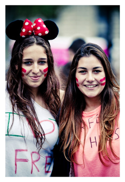 Two young women smiling, one wearing a black Mickey Mouse headband with a red polka-dot bow. Both have red streaks painted on their cheeks and are dressed casually, with "Techno Parade 2013" handwritten on their shirts. The background is blurred, focusing attention on the lively scene at the 15ème édition.
