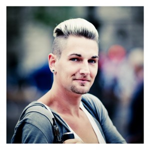 A person with short platinum blonde hair and dark roots, sporting a goatee, pierced eyebrows, and earrings, smiles at the camera while holding an object. The scene appears to be from the Techno Parade 2013. The background is blurred, emphasizing the individual's expressive blue eyes and stylish appearance.