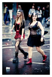 Two women dressed in eclectic, gothic-influenced outfits walk down a wet city street during the 15ème édition of the 2013 Techno Parade. Both have face paint and wear black boots. The woman on the left wears a red plaid skirt and fishnet tights, while the woman on the right sports a black skirt and a lace top.