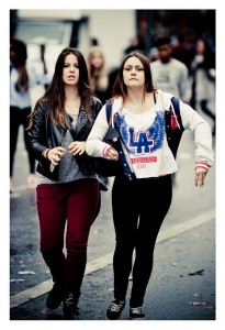 Two young women walk down a street during the Techno Parade's 15ème édition in 2013. One sports a black leather jacket and red pants, while the other wears a white hoodie with blue and red lettering. Both have long hair and are holding onto their bags, surrounded by bustling crowds in the background.