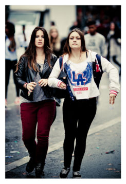 Two young women walk down a street during the Techno Parade's 15ème édition in 2013. One sports a black leather jacket and red pants, while the other wears a white hoodie with blue and red lettering. Both have long hair and are holding onto their bags, surrounded by bustling crowds in the background.