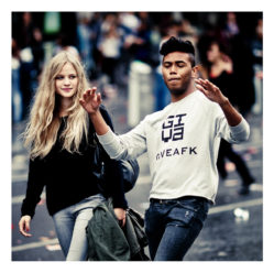 A young woman with long blond hair and a young man with short dark hair are walking together outdoors during the Techno Parade 2013. The woman is smiling and carrying a jacket, while the man is gesturing with his arms and wearing a shirt that says "GIVEAFK.