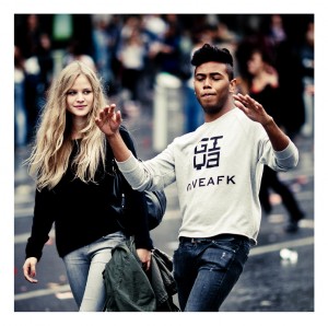 A young woman with long blond hair and a young man with short dark hair are walking together outdoors during the Techno Parade 2013. The woman is smiling and carrying a jacket, while the man is gesturing with his arms and wearing a shirt that says "GIVEAFK.