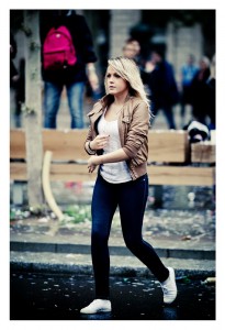A woman with blonde hair wearing a brown jacket, white top, dark jeans, and white shoes is walking on a wet street with a determined look. There are blurred pedestrians and a pink backpack in the background, maybe from the Techno Parade 2013.