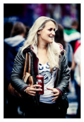 A woman with long blonde hair, wearing a white shirt with a print, black leather jacket, and jeans, stands outdoors smiling at the 15ème édition of the 2013 Techno Parade. She holds a red beverage can in her right hand and has a brown bag over her shoulder. The background shows other blurry people.