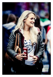 A woman with long blonde hair, wearing a white shirt with a print, black leather jacket, and jeans, stands outdoors smiling at the 15ème édition of the 2013 Techno Parade. She holds a red beverage can in her right hand and has a brown bag over her shoulder. The background shows other blurry people.