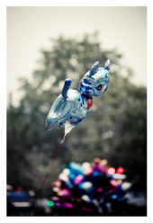 A shiny metallic blue balloon shaped like an animal floats in the air against a blurred background of greenery and colorful balloons, celebrating Techno Parade 2013. The balloon is tethered by a red string, marking the excitement of the 15ème édition.